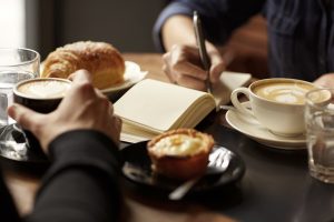 Cropped image of two people's hands at a table with coffees and pastry snacks, one person picking up their espresso while the other is writing in a notebook, possibly taking down an interview