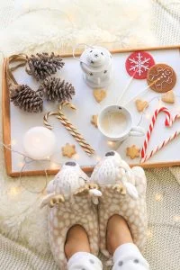Overhead view of girl feet in soft cozy deer slippers and tray with cappuccino, candle, lollipop with ginger man, Christmas candy cane, star cookies, decorative light, pine cones with garland on ivory fur rug and knitted blanket. (Overhead view of gir