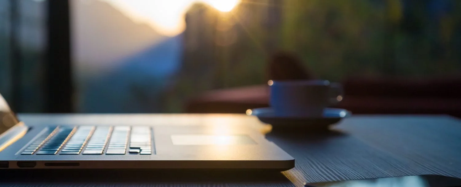 Computer and coffee cup on table. Blurred out mountains in background.