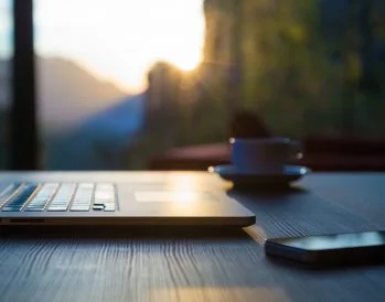 Computer and coffee cup on table. Blurred out mountains in background.