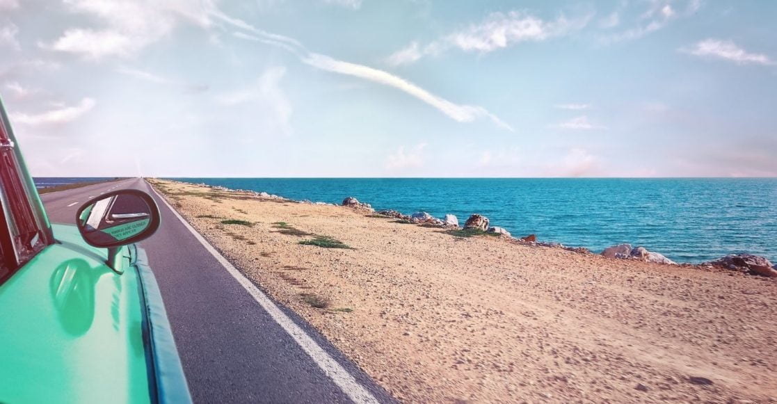 Teal colored car driving down a beachside road with a view of the ocean.
