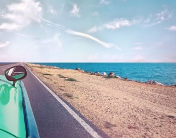 Teal colored car driving down a beachside road with a view of the ocean.
