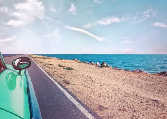 Teal colored car driving down a beachside road with a view of the ocean.